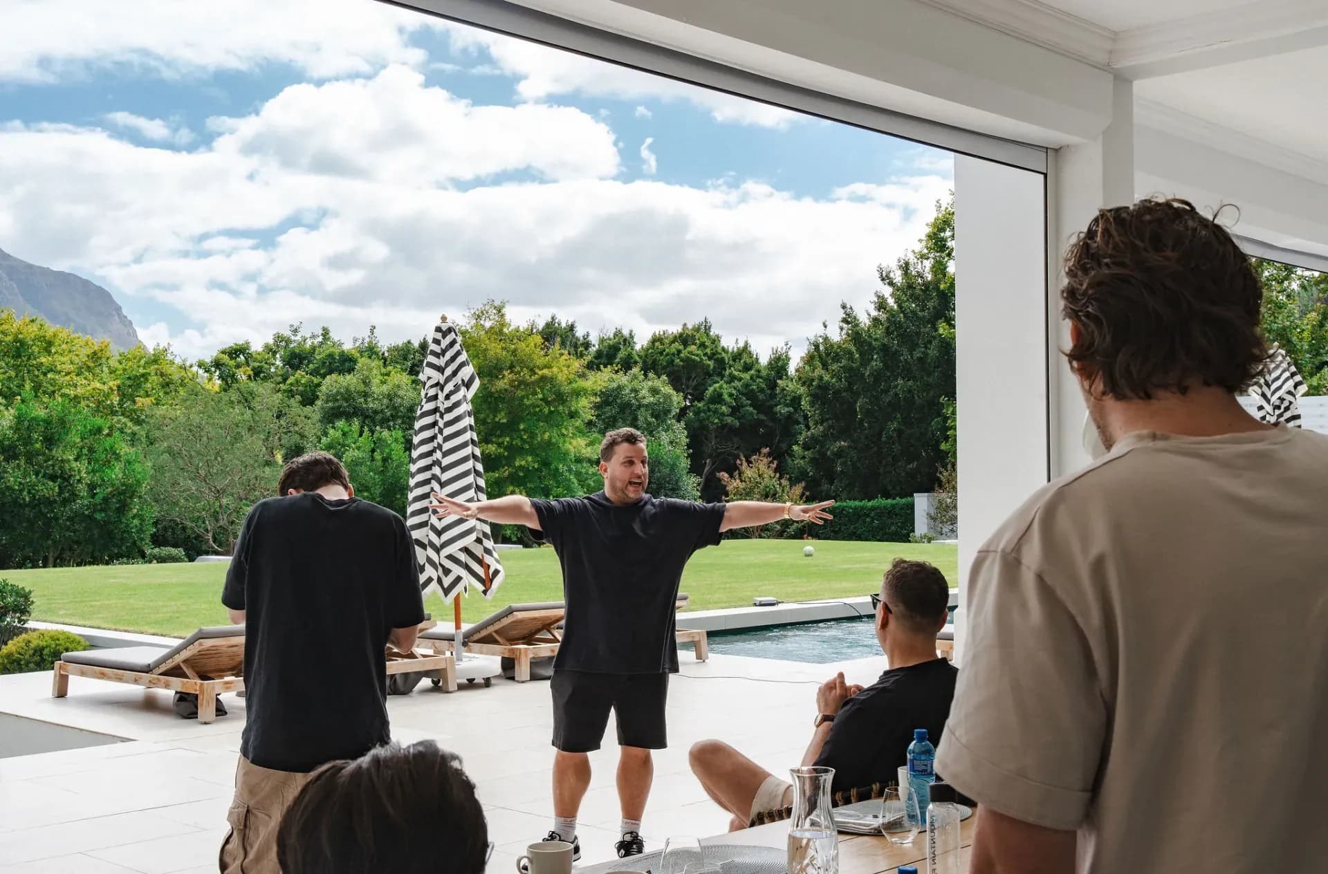 Een founder spreidt zijn armen wijd op het terras van de villa, met de bergen achter hem.