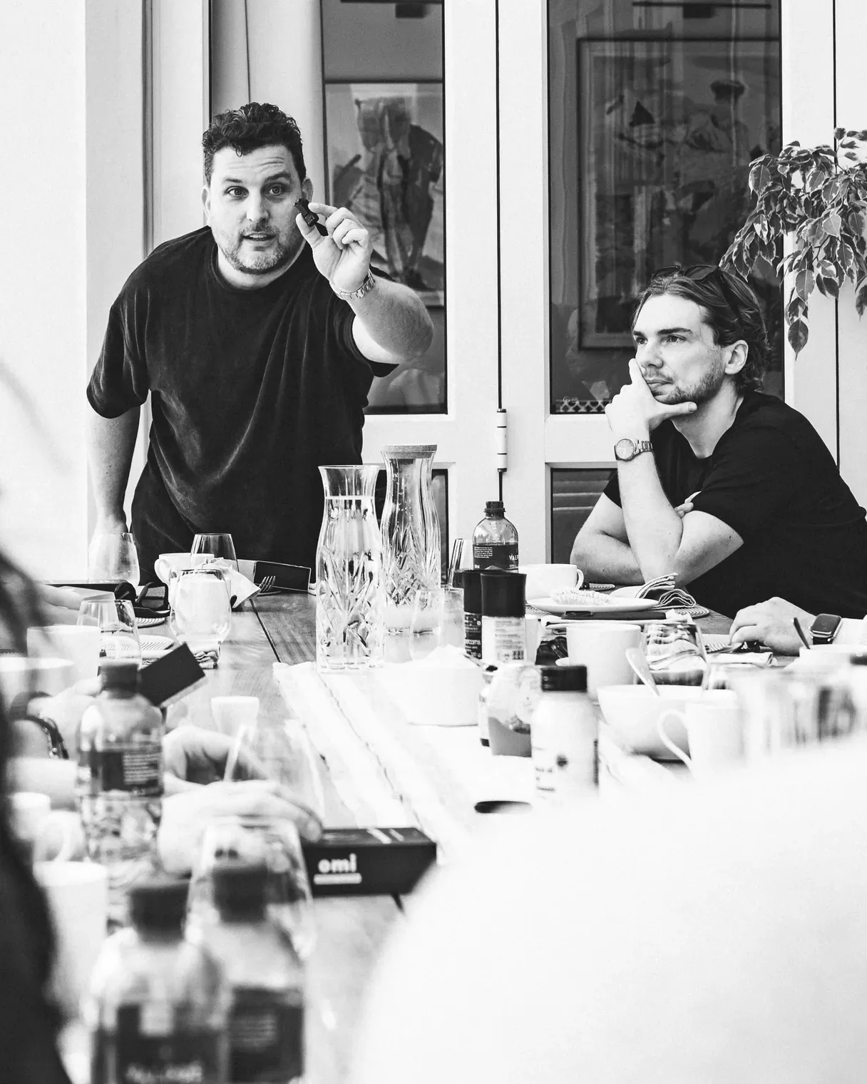 Zwart-witfoto: een founder legt iets uit aan de lange tafel in de villa, terwijl een ander luistert.